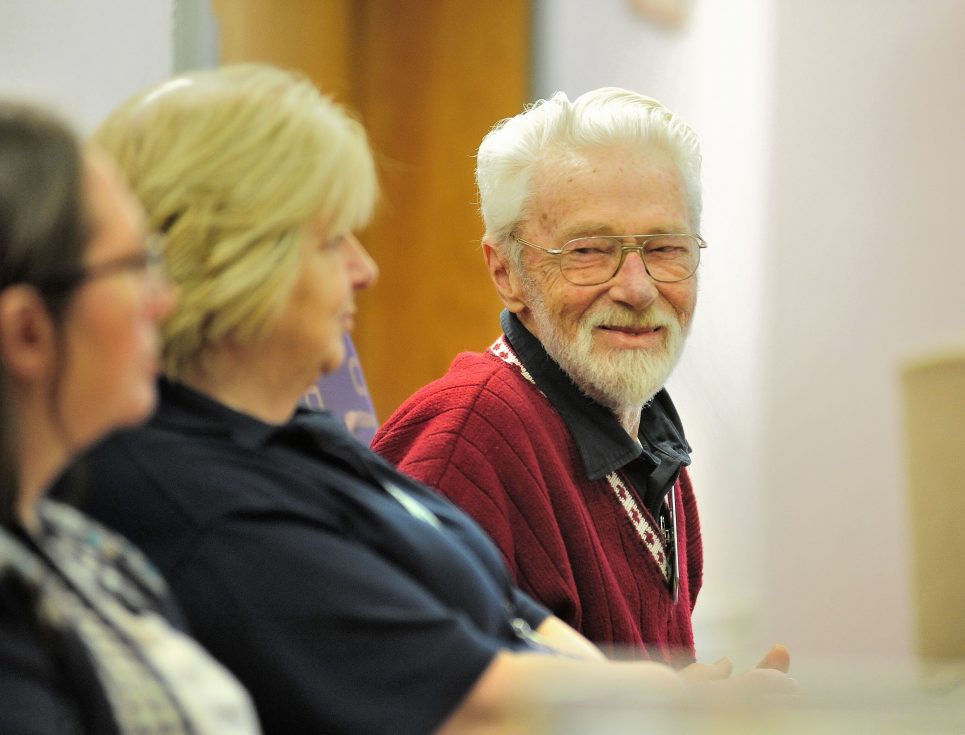 Three people sat in the hospice's Living Well Centre. The camera focuses on a man with white hair, wearing a red jumper and smiling.