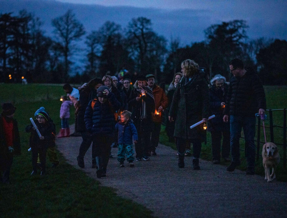 A group of people walk through parkland of Croome Court at twilight. They are holding glowing lanterns and smiling.
