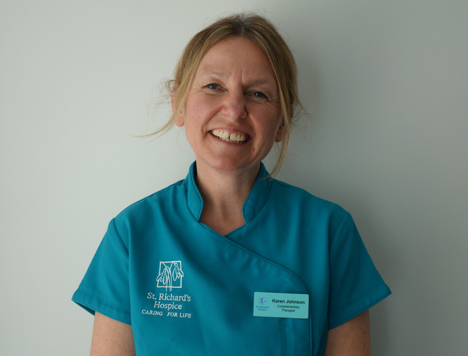 A hospice complementary therapist with her hair tied back is pictured wearing a teal uniform. She is smiling.