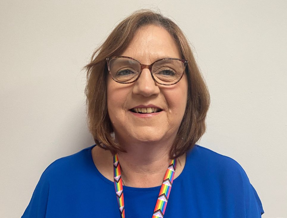 Sue standing in front of a plain white wall. She is wearing a royal blue top and lanyard featuring the progress Pride flag.