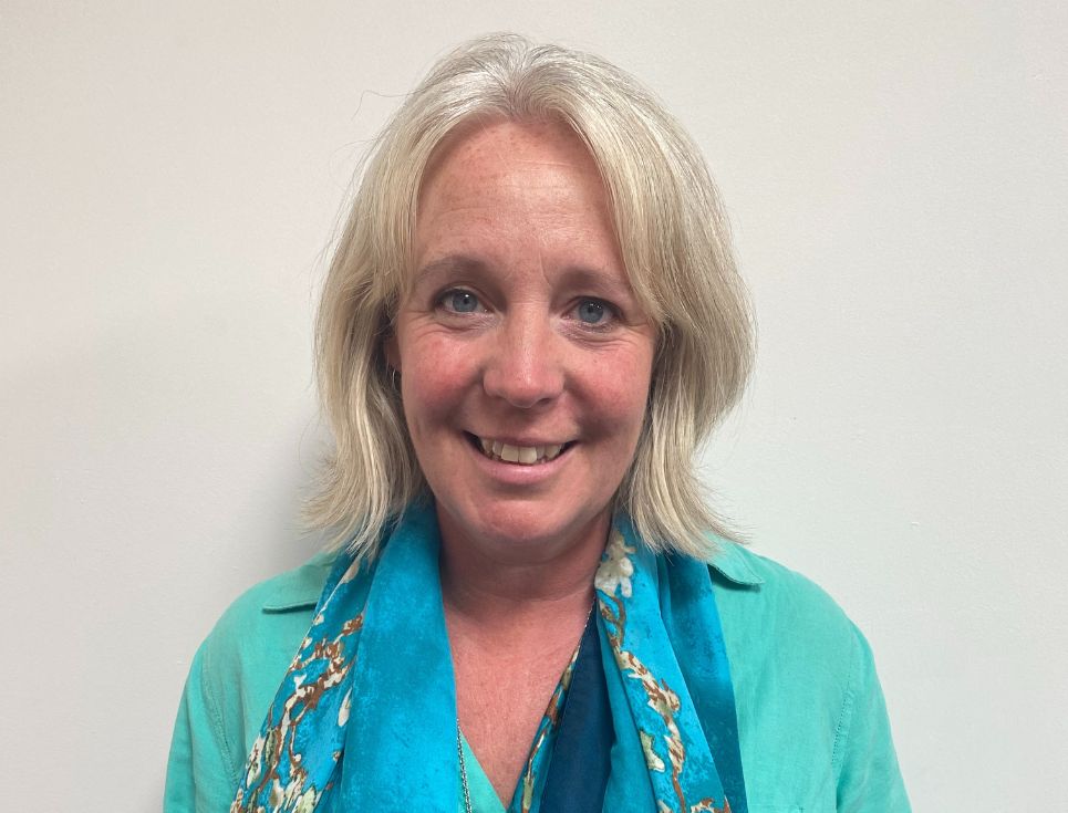 Vicki pictured against a plain white wall. She is wearing a sea green shirt and bright blue scarf. She is smiling.