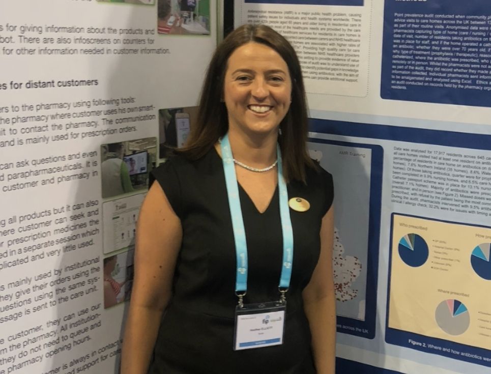 Heather stands between two large posters featuring lots of text and graphs. She is wearing a black dress and has long brown hair. She is smiling.