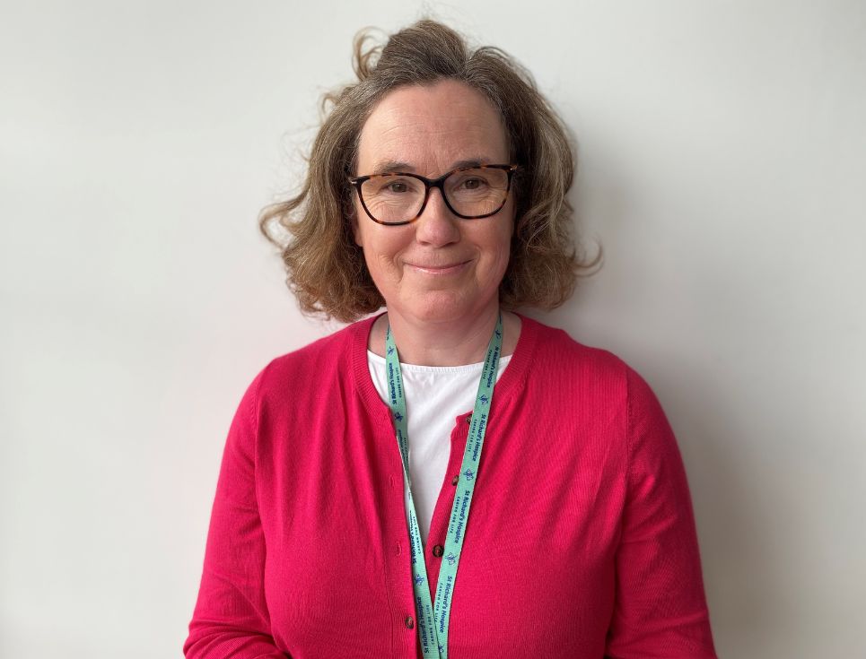 Vicky pictured in front of a plain white wall at the hospice. She is wearing a raspberry pink cardigan buttoned up over a white t-shirt. She is wearing a St Richard's Hospice branded lanyard. She is smiling.