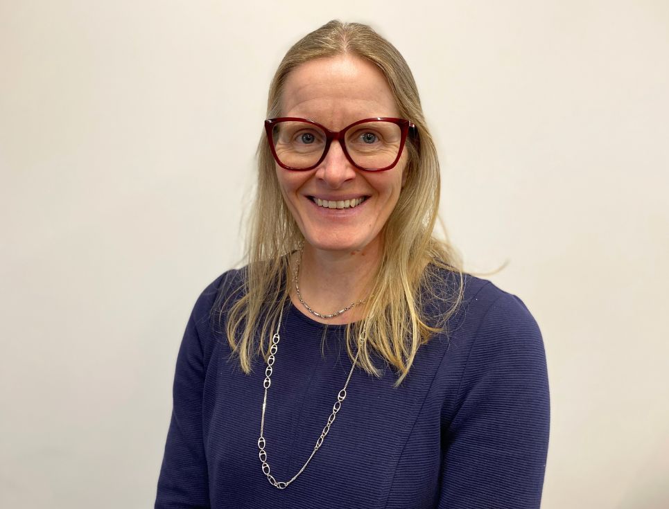 A head and shoulders photo of Jane Nicol with a plain white background behind her. She is smiling and wearing a navy long-sleeved dress. She has shoulder-length blonde hair and wears glasses.