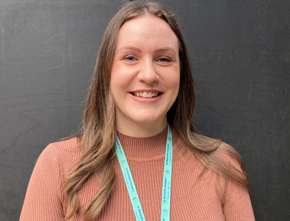 Joanna, pictured in front of a black background. She has long brown hair and is wearing a orange top. She is smiling.