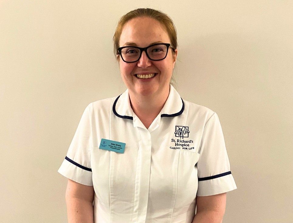 Alex stands in front of a plain white wall. She is smiling and wearing her white physiotherapist's uniform and black framed glasses.