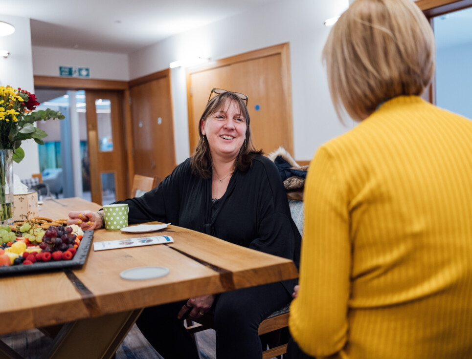 Two people sit talking to each other around an oak table in the Living Well Centre.