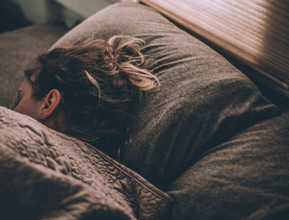 A woman asleep in bed with grey bedding. The woman's head is turned away so you can only see the back of her head.
