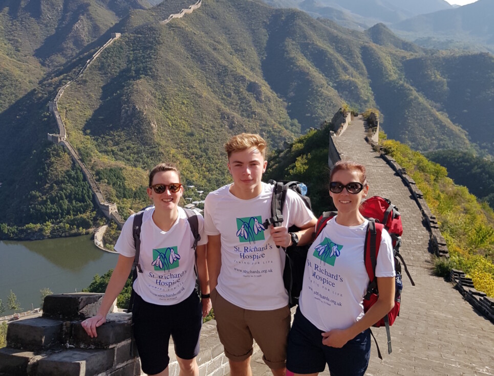 Three people wearing shorts and St Richard's branded t-shirts stand on the Great Wall of China. The wall stretches out behind them, along with rolling hills and blue skies.