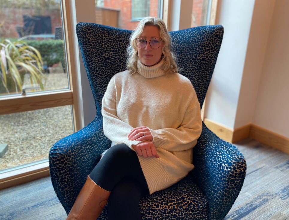 Rebecca sits on a comfy blue, wing-backed chair. She is wearing a cosy, white, roll neck jumper and leggings. She has blonde, shoulder-length hair.