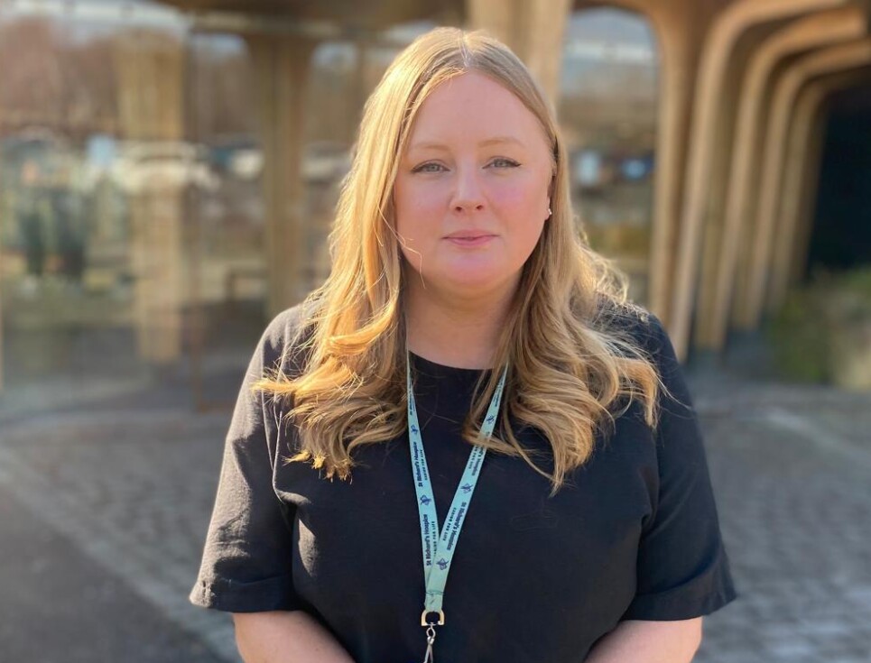 Rebecka Sparks wearing a black top and trousers stands outside the hospice entrance on a sunny day.