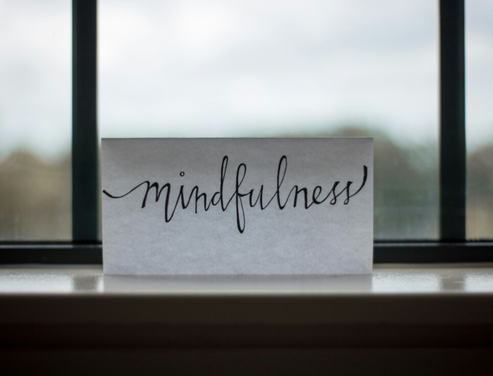 A piece of paper with the word mindfulness written in black ink. Behind the paper are windows.