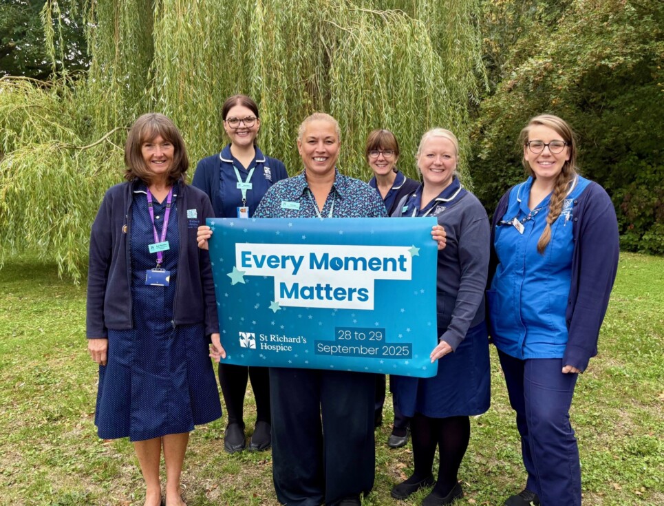 A group of nurses stand together in the hospice gardens holding a banner which reads Every Moment Matters.