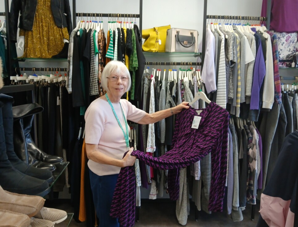 Volunteer Stella stands holding a dress she is about to put on a rail of clothes in the hospice's superstore. She is smiling.